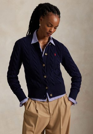 Sötét fonott hajú nő, aki egy tengerészkék fonott mintás kardigánt visel csíkos ing fölött, valamint bézs pliszírozott nadrágot, kezét zsebében tartva, lefelé néz.