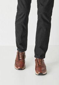 Baskets en cuir marron avec une tige textur�ée, une bande élastique et une semelle de couleur claire, portées avec un pantalon noir.