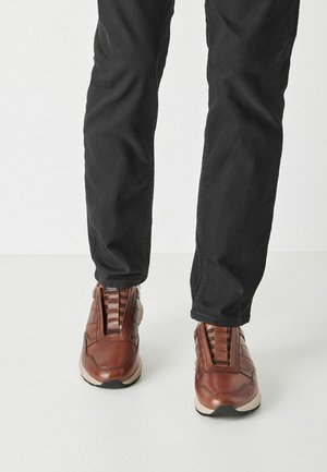 Sneakers in pelle marrone con tomaia testurizzata, fascia elastica e suola di colore chiaro, indossate con pantaloni neri.