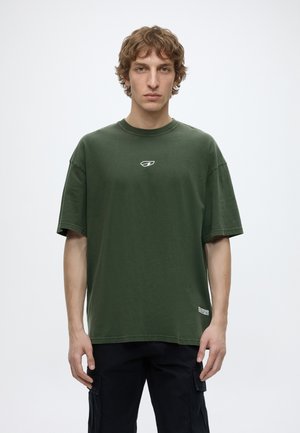 Junger Mann mit lockigem Haar, der ein lockeres dunkelgrünes T-Shirt und schwarze Cargohosen trägt und vor einem schlichten weißen Hintergrund steht.