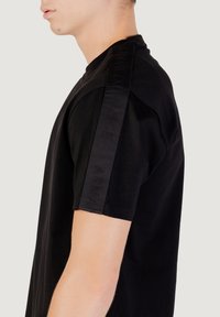 Mustmust t-särk lühikeste varrukatega, millel on ühel õlal tooniline brändi aktsent. Sile tekstuur, standardselt ümmargune kaelus.