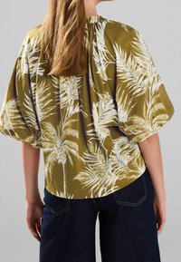 Olive green blouse featuring white palm leaf patterns, short puff sleeves, and a gathered neckline. Paired with dark denim.