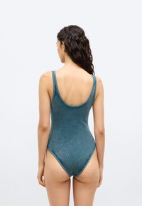 Γυναίκα με μακριά σκούρα σγουρά μαλλιά, φοράει κορμάκι σε σμαραγδί χρώμα με ριγέ υφή και χωρίς μανίκια, εμφανίζεται από πίσω σε απλό φωτεινό φόντο.