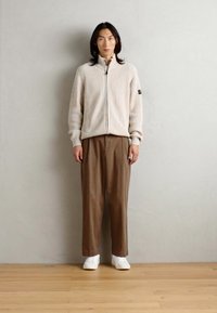 Strikket jakke med glidelås i beige ribbestruktur, kombinert med løse brune bukser og hvite joggesko. Enkel design uten synlige mønstre.