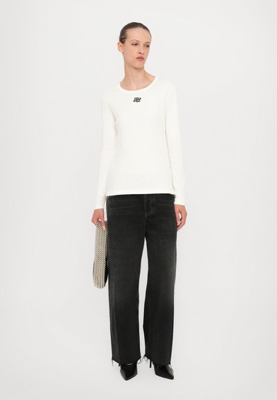 White long-sleeve ribbed top with black logo, paired with high-waisted dark gray wide-leg jeans, and accessorized with a metallic clutch.