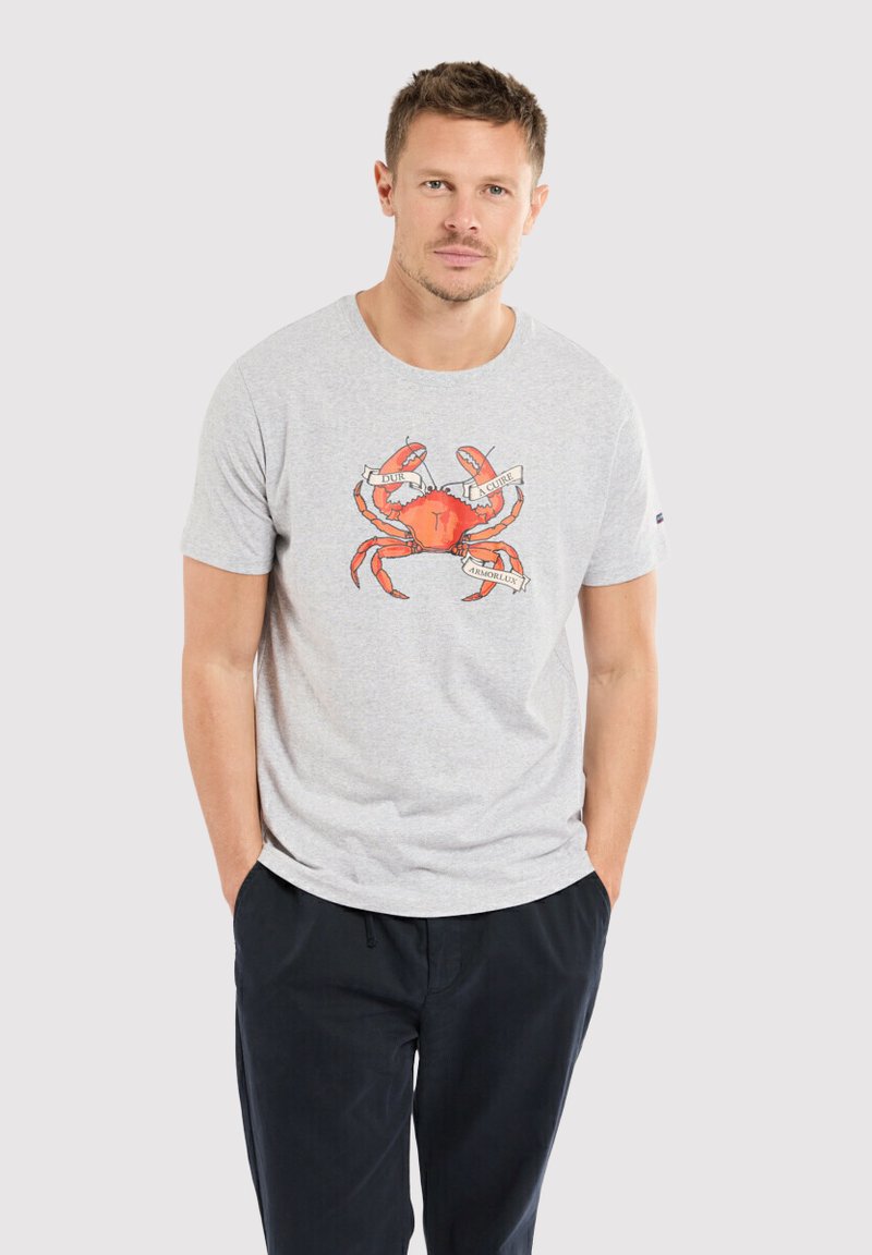 T-shirt en coton gris avec un graphique de crabe rouge au centre, accompagné d'une bannière avec du texte. Le modèle porte un pantalon bleu marine.
