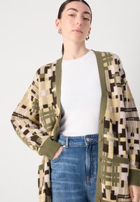 Adīta džemperis ar ģeometrisku rakstu zaļā, bēšā un brūnā krāsā. Pārmērīgi liels dizains ar ribinātām piedurknēm un priekšējiem pogām.