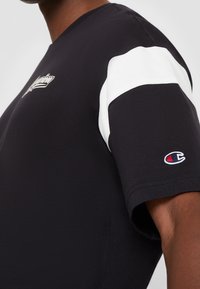 Camiseta negra de manga corta con acento blanco en el hombro, que presenta un logo y una textura suave. Cuello redondo, corte casual.