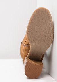 Hnědé semišové kotníkové boty s texturovanou gumovou podešví a krátkým, vrstveným podpatkem. Disponují bočním zipem pro snadné obouvání.