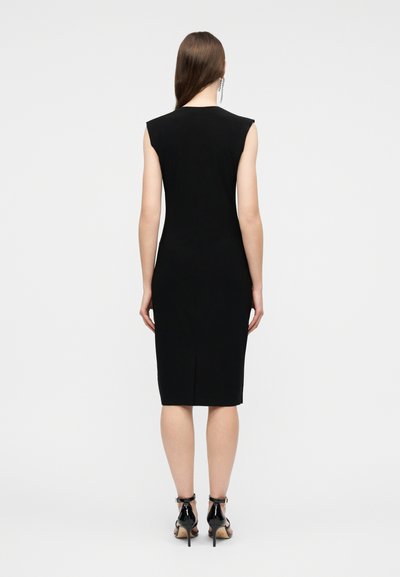 Γυναίκα που φοράει μαύρο, αμάνικο φόρεμα μέχρι τα γόνατα και μαύρα σανδάλια με ψηλό τακούνι, στέκεται πλάτη σε ένα απλό φόντο.