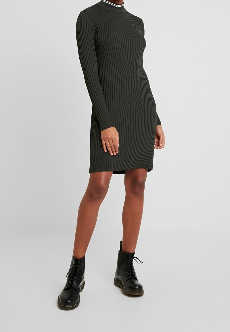 Robe moulante à côtes de couleur vert foncé avec manches longues et col montant, ornée d'un col rayé blanc. Portée avec des bottes noires à lacets.