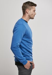 Zils džemperis no mīkstas auduma, ar apaļu kakla izgriezumu, ribotām piedurknēm un apakšmalu. Modelis uzvilcis pelēkas bikses, skatīts no sāniem uz neitrāla fona.