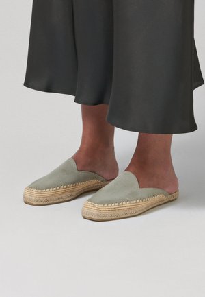 Nohy v světlých zelených nazouvacích espadrillových mulech s pletenými jutovými podešvemi, spárované s širokými černými zkrácenými kalhotami.