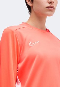 Крупен план на човек, носещ спортна риза с дълги ръкави Nike в коралов цвят с бяло лого под формата на крила на гърдите.
