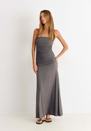Frau mit langen Haaren, die ein trägerloses graues Maxikleid mit Raffung und schwarzen Sandalen trägt, steht vor einem schlichten weißen Hintergrund.