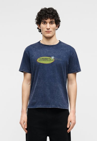 Νεαρός άνδρας με σγουρά σκουρόχρωμα μαλλιά φοράει ένα ξεθωριασμένο μπλε ναυτικό μπλουζάκι με το λογότυπο "MISBHV clubwear solutions", στέκεται μπροστά σε ένα απλό φωτεινό φόντο.