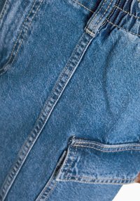 Džínové kalhoty ve středně modré barvě, s nařaseným pasem, viditelným šitím a boční kapsou pro lepší funkčnost.