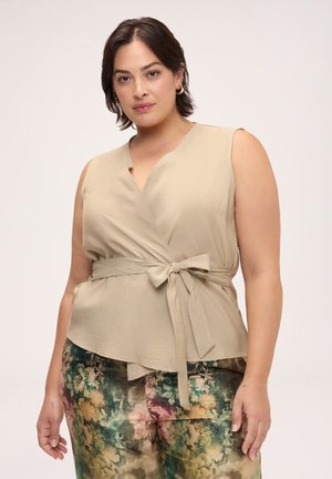 Donna che indossa un top beige senza maniche con incrocio e vita annodata e pantaloni con fantasia floreale, in piedi davanti a uno sfondo chiaro uniforme.