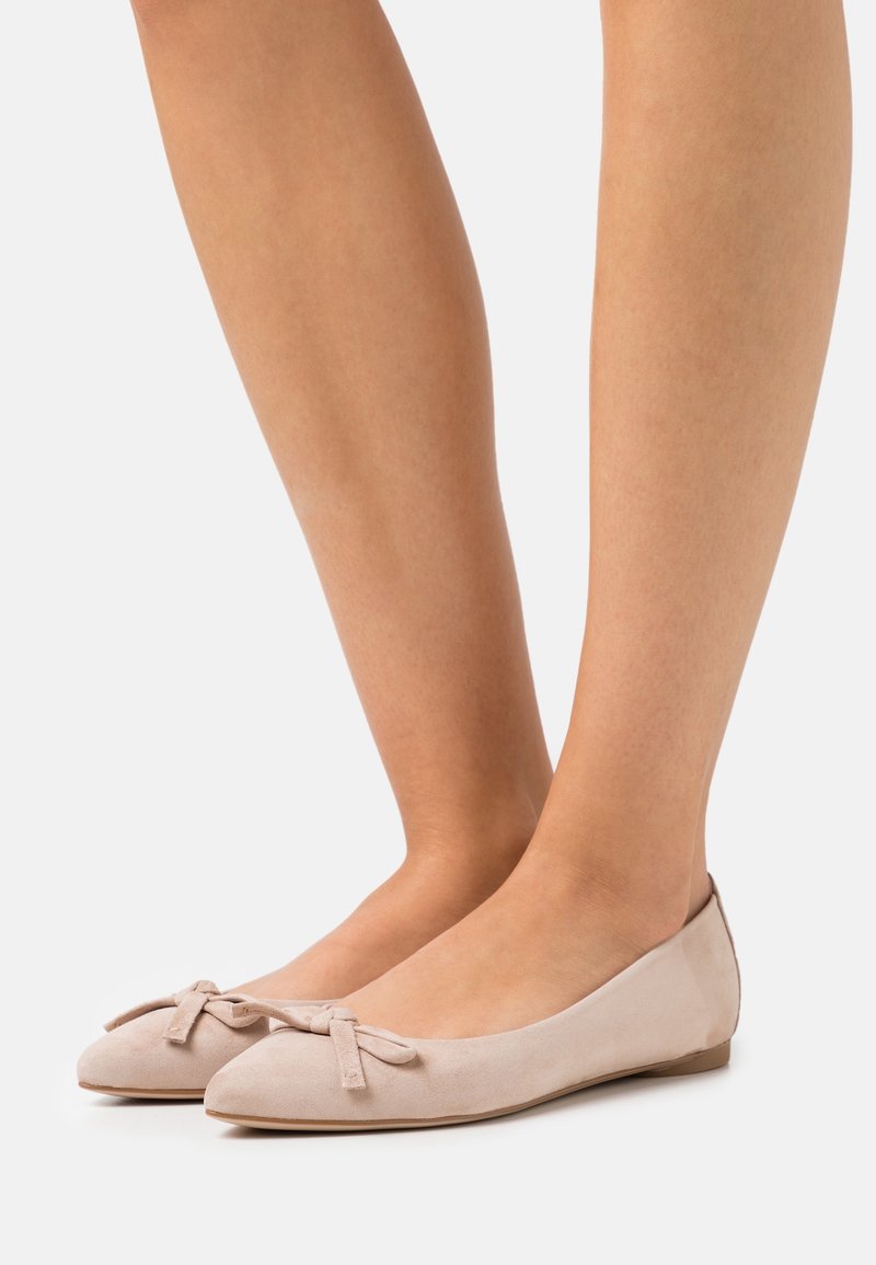 Pieds portant des ballerines en daim beige clair avec un détail nœud sur le dessus, posés sur un fond blanc uni.