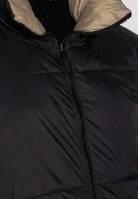 Czarna kurtka puchowa z beżowym wnętrzem, z wysokim kołnierzem, pełnym zamkiem z przodu i pikowanym wzorem zapewniającym izolację.