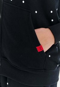 Czarna bluza z białymi kropkami, z przodu na zamek błyskawiczny i kieszenią z czerwoną metką. Miękka tkanina z ściągaczami przy rękawach i u dołu.