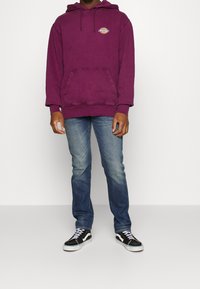 Sweat à capuche en coton violet avec une poche avant et un logo ; associé à un jean en denim bleu clair et des baskets noires avec des accents blancs.