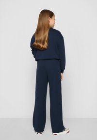 Jūras zilas krāsas sporta krekls un platā kāju bikses no mīksta materiāla. Sporta krekliņam ir brīvs piegriezums; biksēm ir elastīga josta un smalkā šuvuma detaļas.