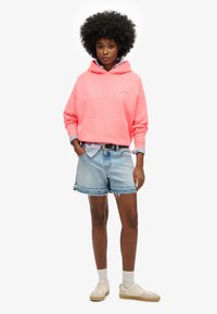 Haut à capuche rose avec une poche avant, associé à un short en jean bleu clair. Le modèle porte des baskets crème et des chaussettes blanches à la cheville.