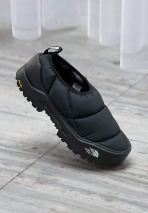 Black slip-on shoe with a puffy, quilted upper, textured rubber sole, and distinctive logo. Designed for comfort and casual wear.