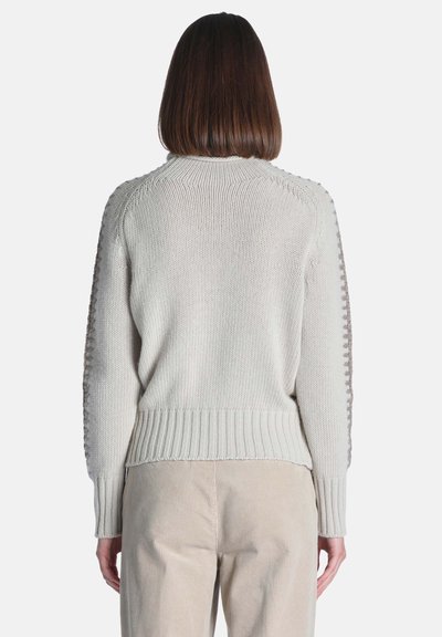 Femme aux cheveux châtain mi-longs portant un pull en maille crème et un pantalon beige, vue de dos sur un fond uni.