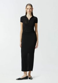 Crna polo haljina s uskim siluetom, kratkim rukavima, dugmetima i bočnim prorezom. Nosena s crnim ravnim cipelama.