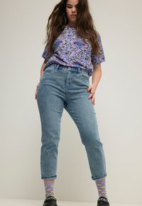 Vaaleansiniset korkeavyötäröiset farkut, joissa on rento istuvuus, yhdistettynä violettiin kuvioituun paitaan ja keltaisiin hymyileviin sukkiin.