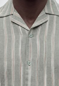 Κοντινό πλάνο ενός ατόμου που φοράει πουκάμισο με υφή και κουμπιά, με κάθετες ρίγες σε γκρι, λευκό και ανοιχτό πορτοκαλί χρώμα και γιακά με χαραγμένο άνοιγμα.