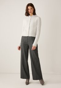 Weißer Cardigan aus Wollmischung mit geripptem Saum und langen Ärmeln, kombiniert mit grauen Hosen mit weitem Bein und taupefarbenen Stiefeletten. Sichtbarer gestreifter Hemdkragen.