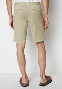 Beige cotton shorts with a straight cut, two back pockets with buttons, and a folded hem. Worn with brown slide sandals.