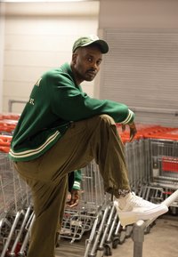 Grön varsityjacka med vita ränder, olivgröna cargobyxor och vita sneakers. Modellen poserar på ett räcke med shoppingvagnar i bakgrunden.