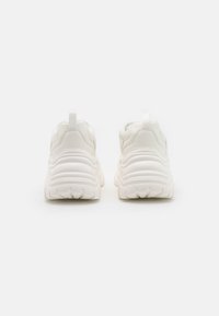 Bílé sportovní boty s tlustou, vroubkovanou podrážkou a hladkým svrškem. Obsahují zadní poutko a strukturované detaily na patě.