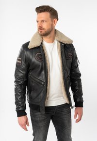 Schwarze Leder-Bomberjacke mit beigem Lammfellkragen, zwei Vorderaschen und gestickten Patches an den Ärmeln. Reißverschluss und gestrickte Bündchen.