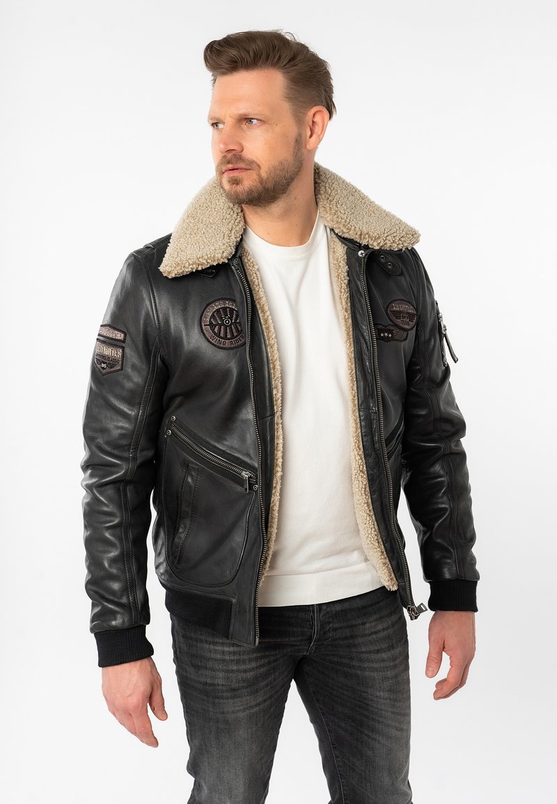 Schwarze Leder-Bomberjacke mit beigem Lammfellkragen, zwei Vorderaschen und gestickten Patches an den Ärmeln. Reißverschluss und gestrickte Bündchen.