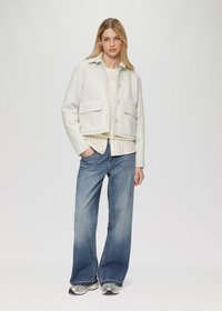 Veste grise claire coupée avec de grandes poches avant, associée à un pull crème et une chemise rayée en dessous, portée avec un jean bleu à jambes larges.