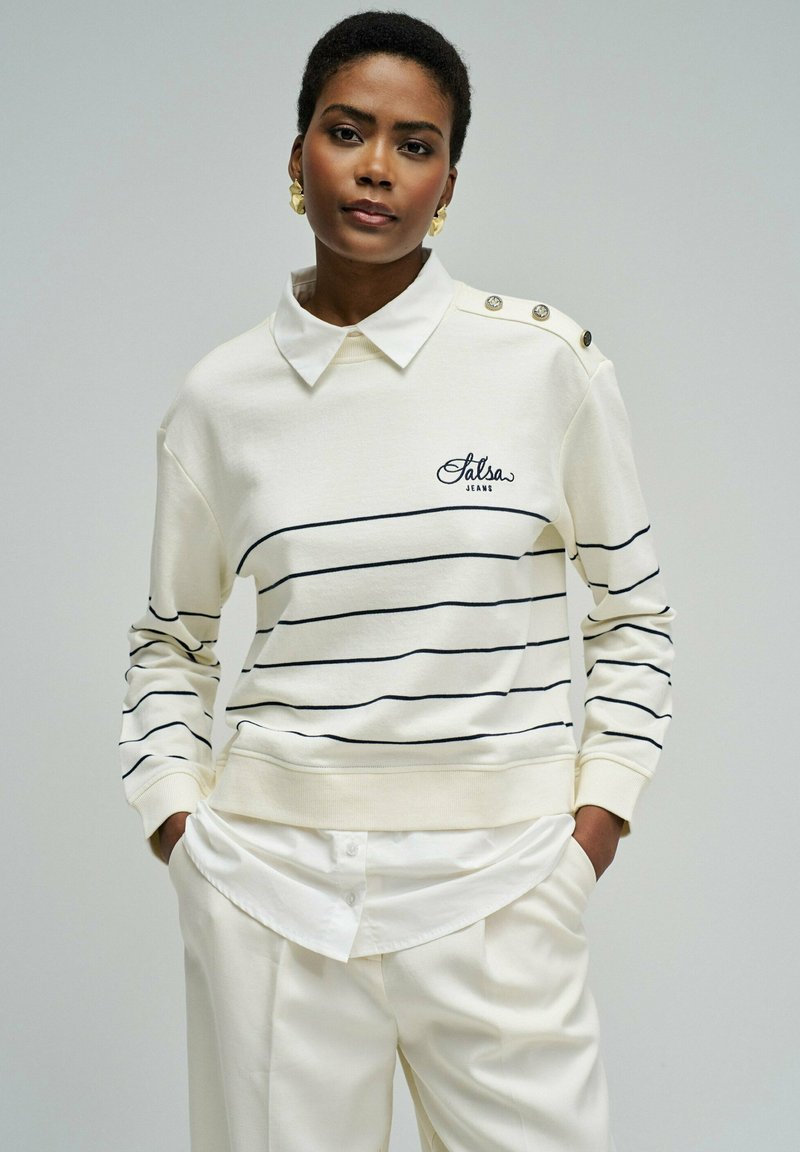 Mujer con un suéter crema con finas rayas negras y botones en el hombro, camisa blanca con cuello y pantalones crema con las manos en los bolsillos.