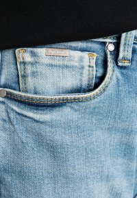Jeans de ganga azul claro com uma textura desgastada, apresentando um botão prateado e uma pequena etiqueta "Pepe Jeans" no bolsinho da frente.