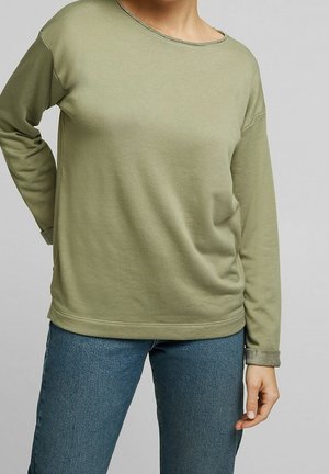 Personne portant une chemise vert clair à manches longues et un jean bleu, debout avec une main sur la hanche et l’autre main détendue le long du corps.