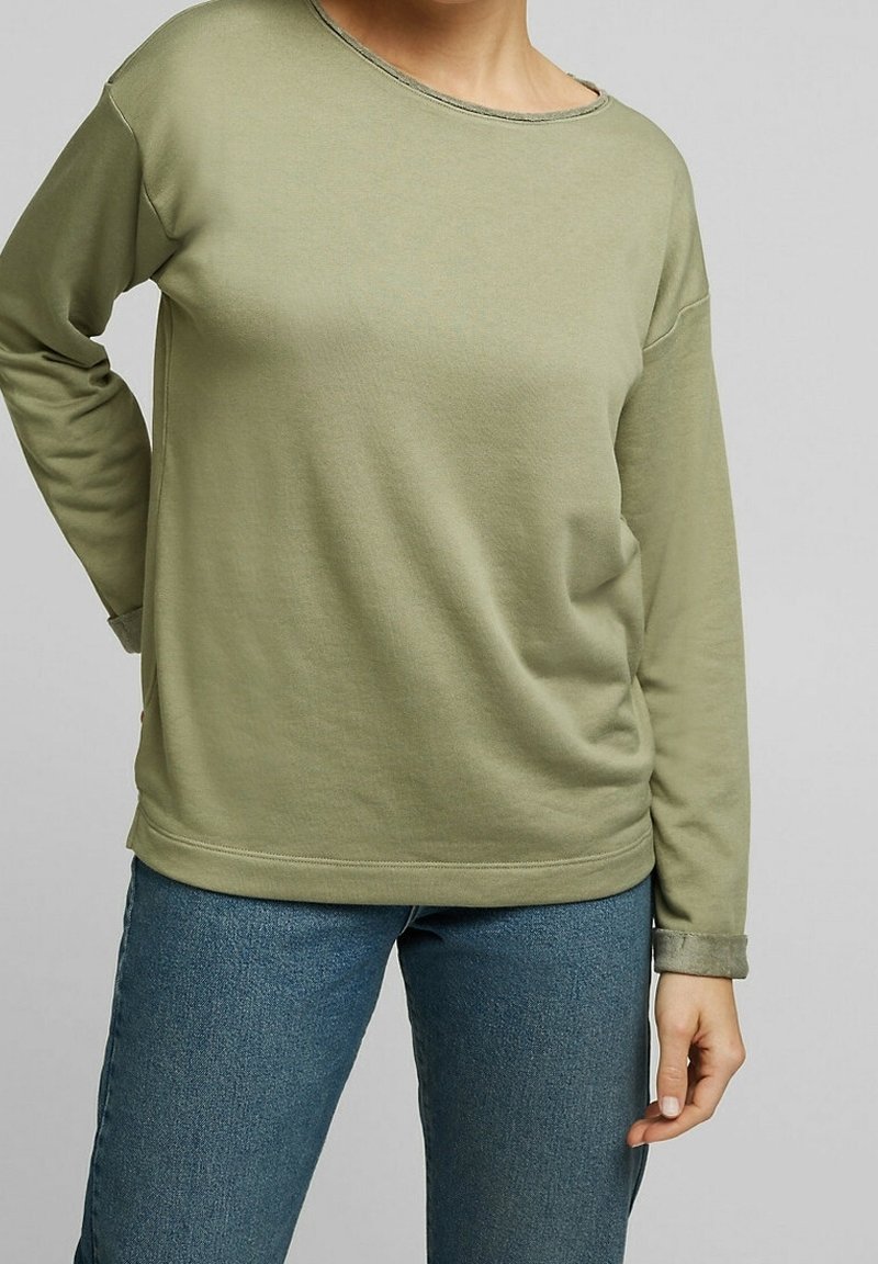 Personne portant une chemise vert clair à manches longues et un jean bleu, debout avec une main sur la hanche et l’autre main détendue le long du corps.