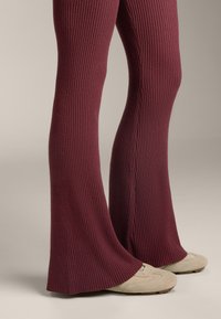 Leggings a coste color marrone borgogna con forma svasata, caratterizzati da un tessuto testurizzato e un dettaglio sulla cucitura laterale, abbinati a scarpe ballet flat di colore chiaro.