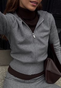 Szara bluzka z suwakiem, z prążkowanymi mankietami i dołem, nałożona na brązowy golf. Brązowa torebka z wyraźnym, kanciastym wzorem widoczna.