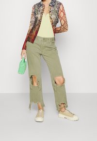 Mujer vestida con pantalones verdes desgastados, zapatillas beige, una blusa de manga larga con estampado de serpiente, una camiseta amarilla y sosteniendo un pequeño bolso verde.