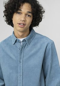 Chemise en velours côtelé bleu clair avec patte de boutonnage à l'avant et col classique. Présente des rayures verticales subtiles et une coupe décontractée.