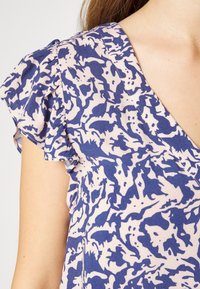 Top com padrão em roxo e bege, com decote em V e mangas curtas com babados. O tecido parece leve e texturizado, com um design ousado.