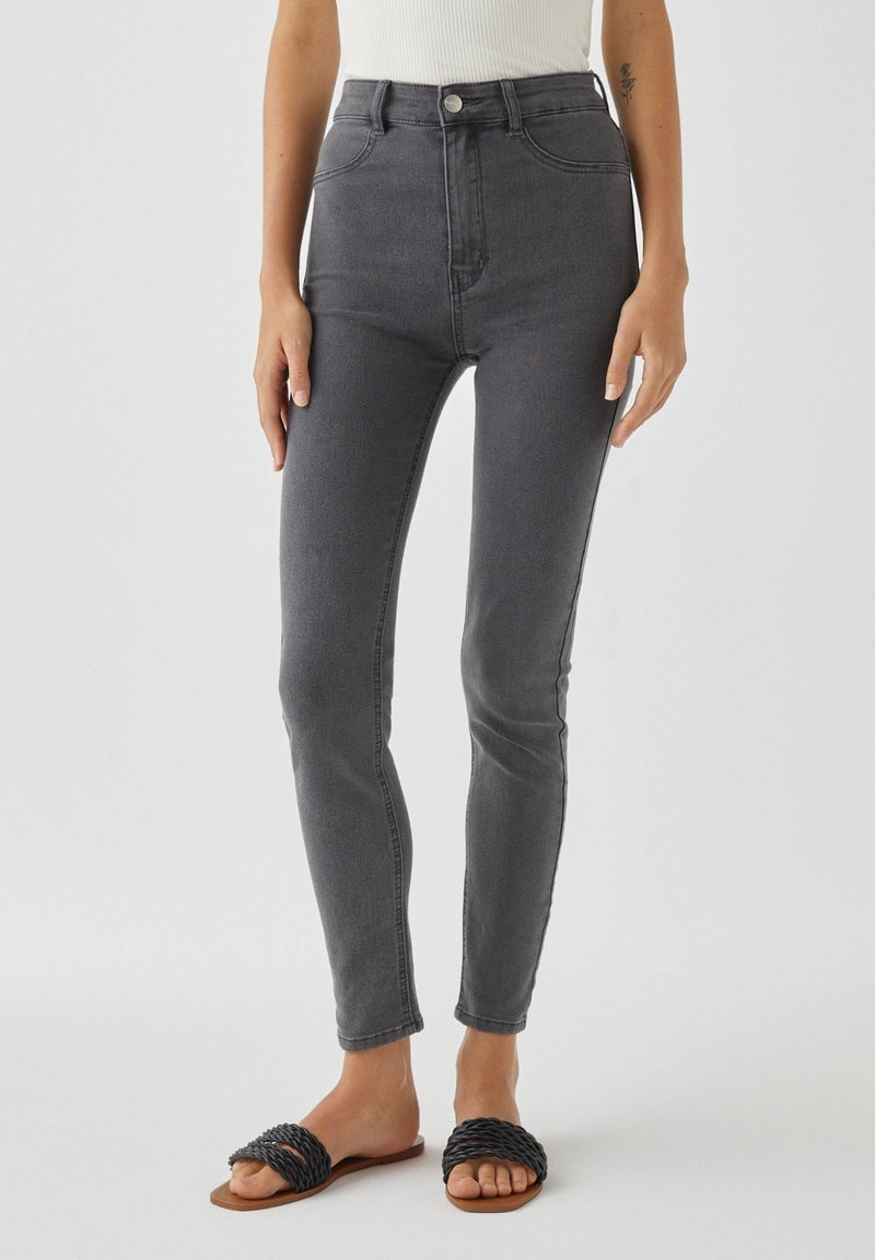 Jean gris taille haute avec une coupe slim, en denim. Comprend des poches avant et une fermeture à bouton. Associé à des sandales à glissière tissées noires.